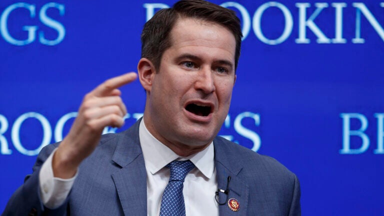 Rep. Seth Moulton, a middle-aged white man with short brown hair, is shown mid-speech, his hand raised in a pointing gesture. He is wearing a blue suit and blue tie.