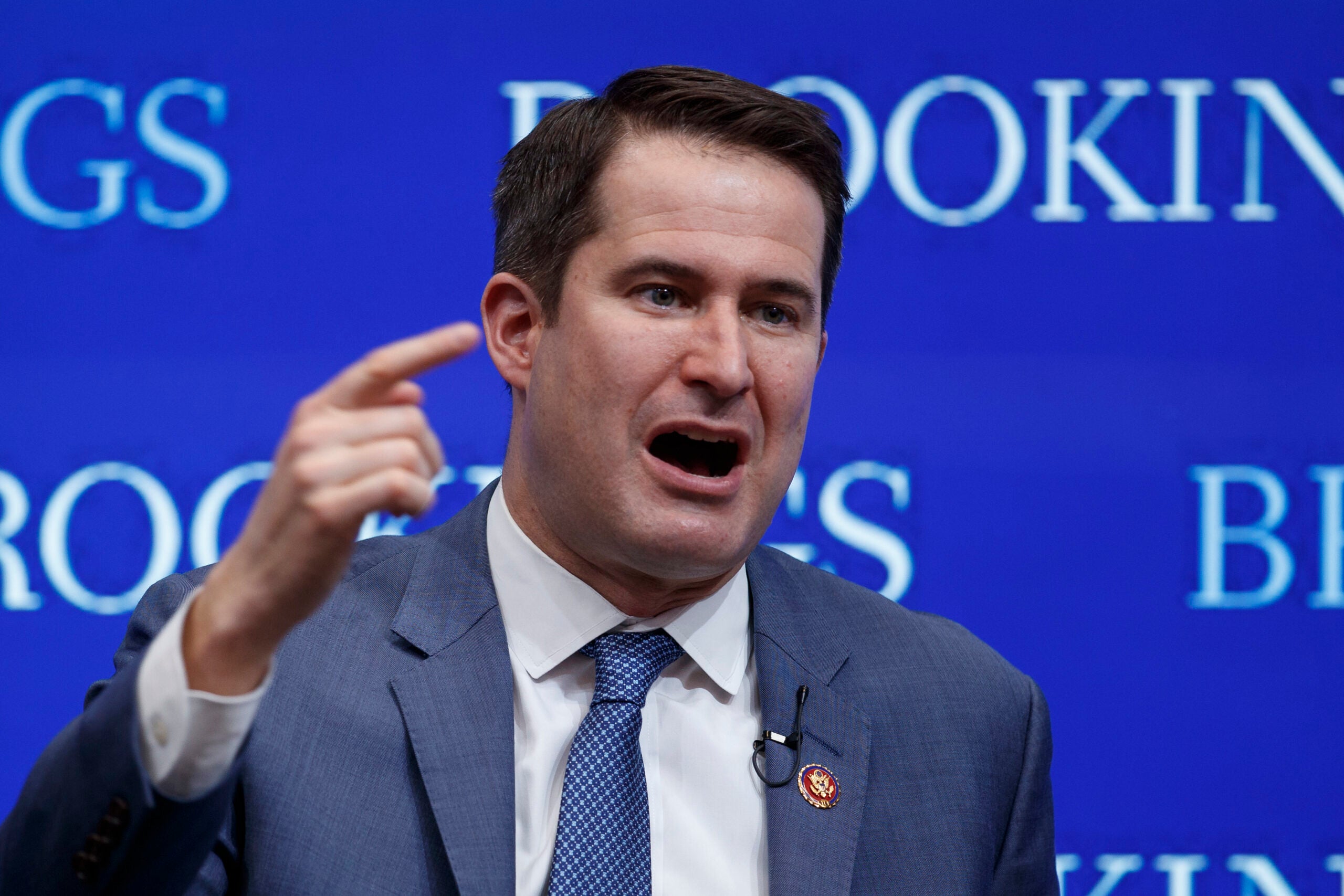 Rep. Seth Moulton, a middle-aged white man with short brown hair, is shown mid-speech, his hand raised in a pointing gesture. He is wearing a blue suit and blue tie.