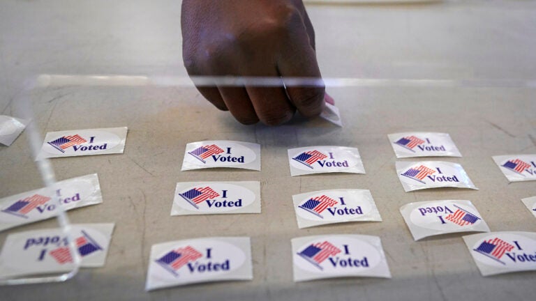 alt = a voter's hand reaches out to grab a white "I voted" sticker from a table of them. The stickers feature an American flag.