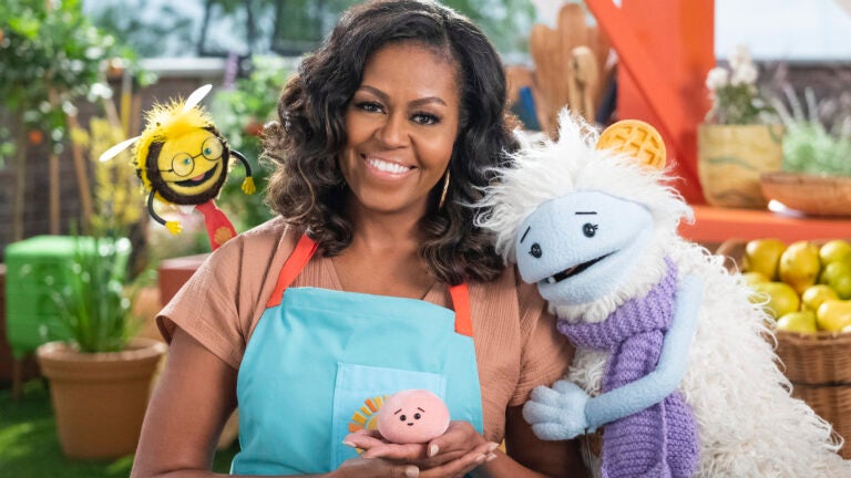 Michelle Obama, wearing a blue apron, stands in a rooftop garden posing for the camera and holding a pink, round mochi puppet. A bee puppet wearing a red tie hovers over her shoulder while a furry white and blue puppet with frozen waffle ears embraces her side.