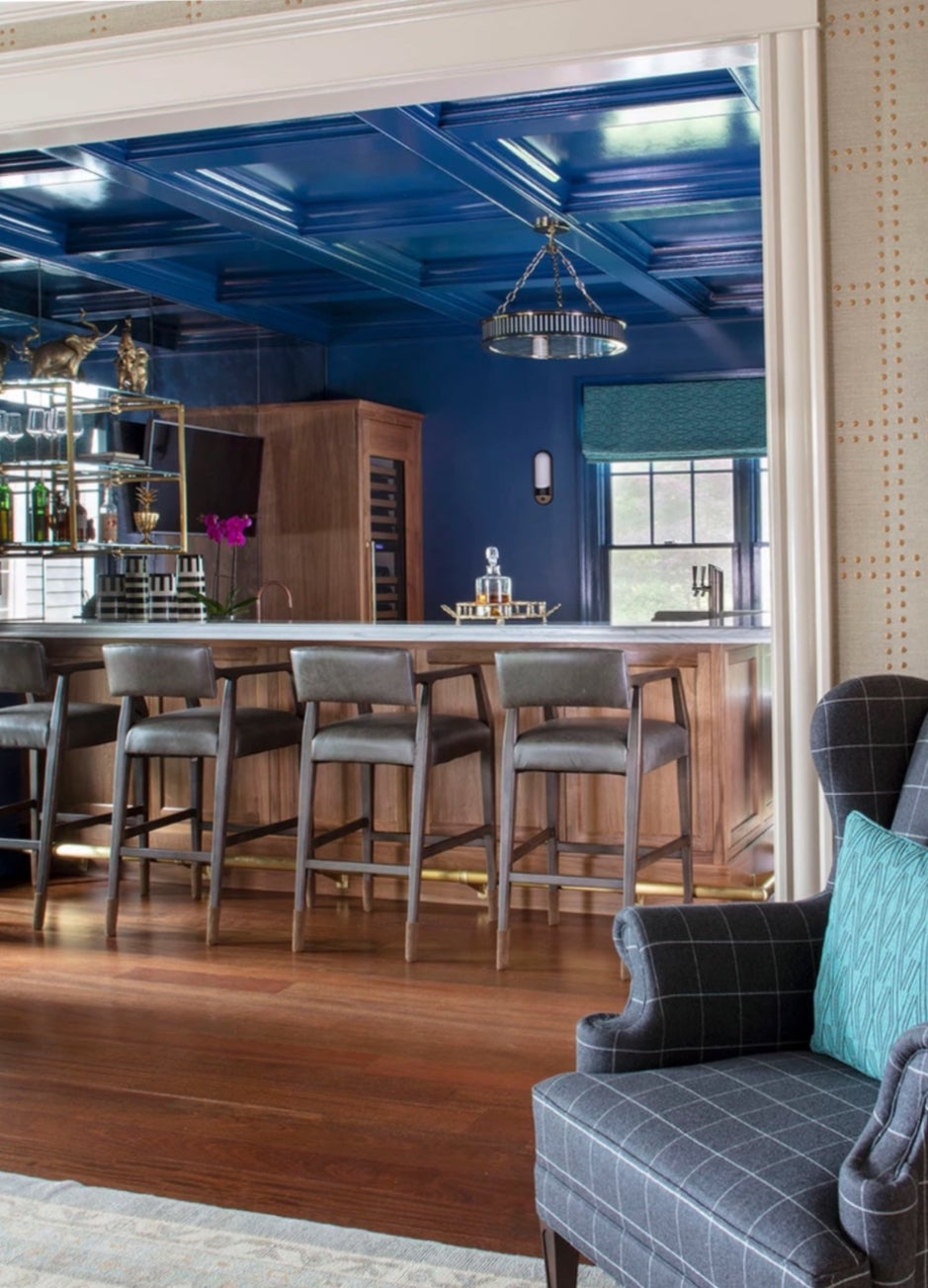 The coffered ceiling and the walls in this space are painted a moody dark blue. There is a wing chair in the foreground with a blue checkered print and a teal, patterned pillow. The bar is in the back. It has a white counter, four hard-backed metal stools, and a brass foot rail. There is a window behind the bar, and the sunlight is bouncing off the glass backsplash behind open shelving filled with liquor bottles. There is a carafe on top of the bar.