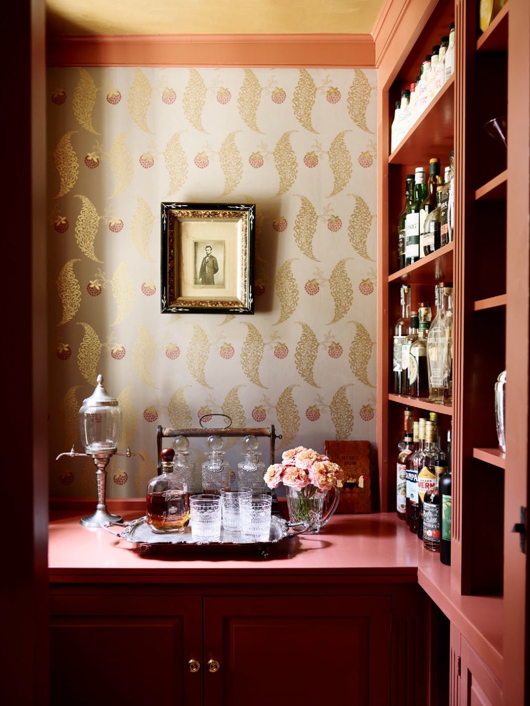 Red bar and cabinetry. The red counter has a silver tray with bar glasses and a round bottle of liquor. There is a short vase of pink flowers. A lantern on a stand is to the left. The wallpaper is gold leaves and red berries. The side shelves are full of liquor bottles.