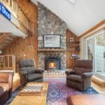A spacious room with high ceilings has a brown leather couch and two cushioned reclining chairs. There’s a decorative carpet on the floor, and a pellet stove is set up against the fireplace, which has a stone backdrop. The floors and back wall are made of wood.