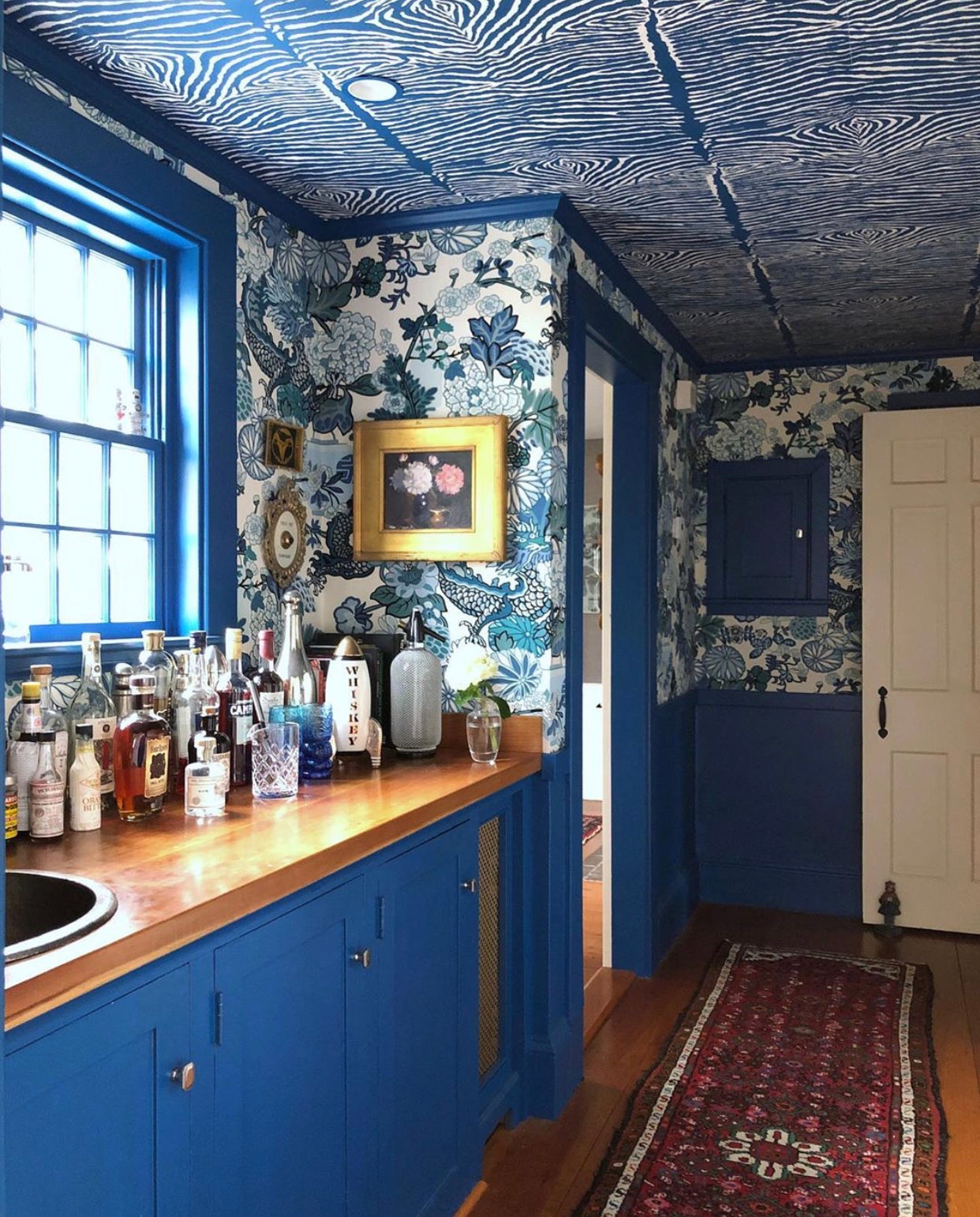 A polyurethaned counter sits atop bright blue cabinetry. Several bottles of liquor sit on top of the counter, glinting in the sunshine spilling in from the blue-framed window above. There is a red, patterned rectangular rug on the floor. The walls are paper in a big-patterned floral paper.