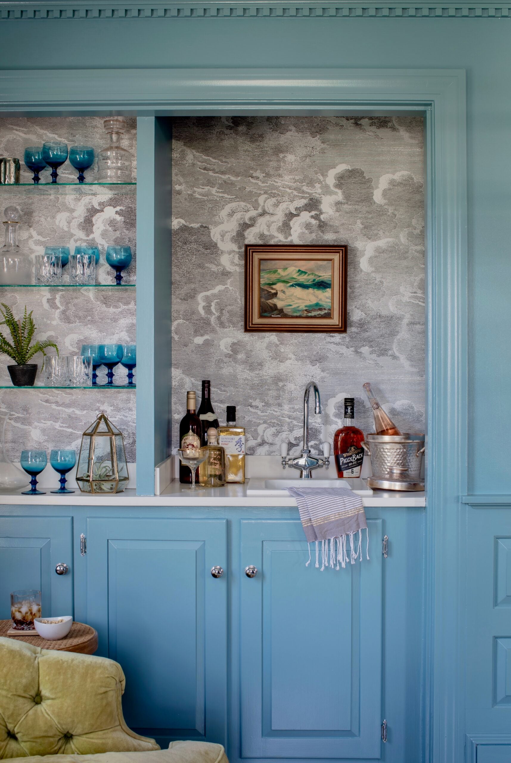 Light blue cabinetry makes up the bar in this space and matches the wainscoting and framing of the room. A fringed tea towel sits in a small sink with a metal faucet. An olive green armchair is in the foreground next to a small round end table with a bowl and a full glass with ice on it. There are blue glasses on the glass open shelves of the bar. The backsplash is a wallpaper with a gray cloud print. There are bottles of liquor and an ice bucket with a bottle in it on the counter.