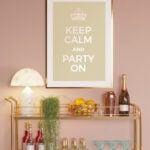 A gold-toned two-tier bar cart is positioned in front of a pink wall and under a sign that reads: Keep calm and party on." There is a white lamp with a round shade on the top shelf, as well as a trailing plant, a basket of fruit, a red shaker, and a bottle of champagne. The second shelf holds four red bottles, six bottles, and rows of champagne glasses.