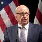 Rupert Murdoch stands before a microphone wearing a dark gray suit, a purple patterned tie, and brown glasses. Two American flags hang in the background.