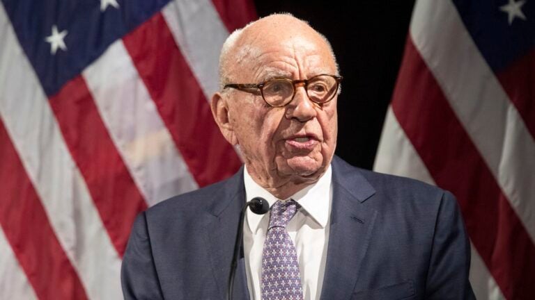 Rupert Murdoch stands before a microphone wearing a dark gray suit, a purple patterned tie, and brown glasses. Two American flags hang in the background.
