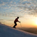 Two skiers head downhill with sun and clouds in the background.