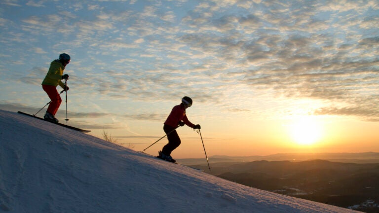 Two skiers head downhill with sun and clouds in the background.