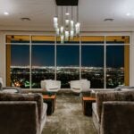 A carpeted room has two wide, comfortable chairs facing each other in the foreground, and two white, modern-looking chairs by the floor-to-ceiling window that looks out over the LA cityscape. A modern light fixture hangs from the ceiling in the center of the room.