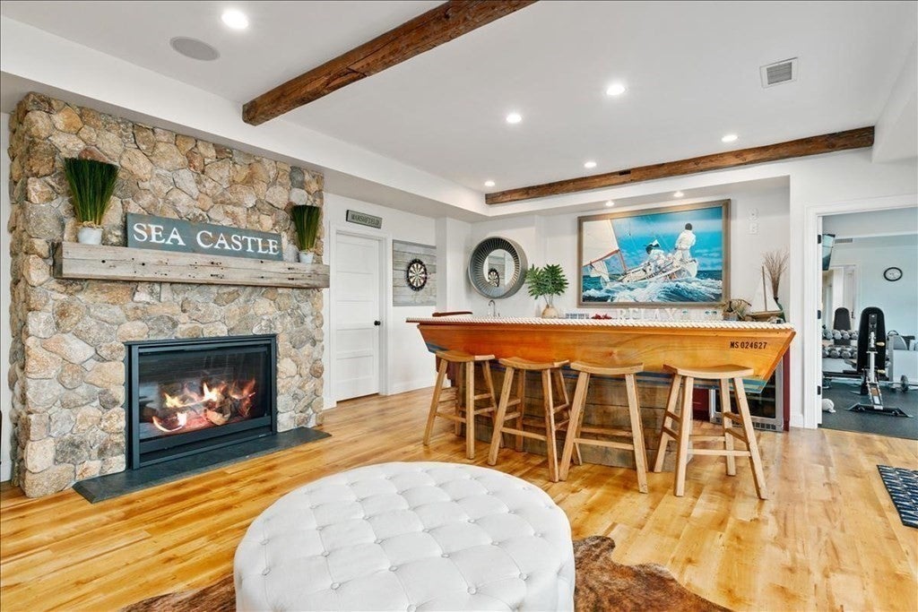 There is puffy white stole in the foreground of this photo of the basement bar. On the left wall, a fireplaces is surrounded by a brick wall. The bar, which is made out of a boat, is located by the back wall. The floors are wood and two wood beams run across the ceiling.
