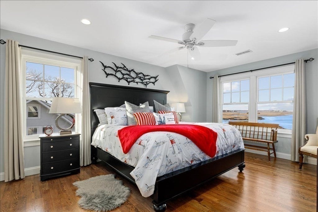A large bed with a black frame and headboard sits against the back wall next to a window. There are two more windows on the right wall, through which the marsh is visible. A ceiling fan hangs in the center of room over the bed.
