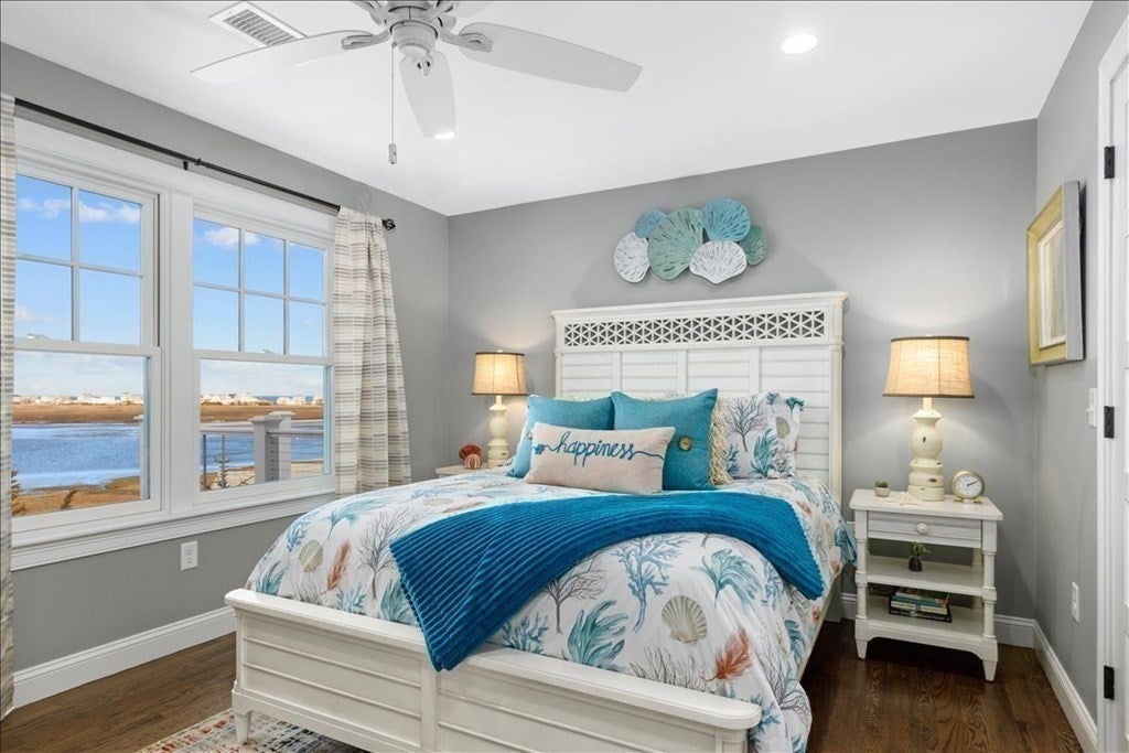 A double bed is set up in the middle of the room, with a short white bedside table next to it on the right. There are two windows that look out to the water on the left wall.