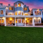 The view of the back of the home at sunset. Light is pouring out of the numerous windows of the three-story structure. There's a green, manicured lawn in the foreground.