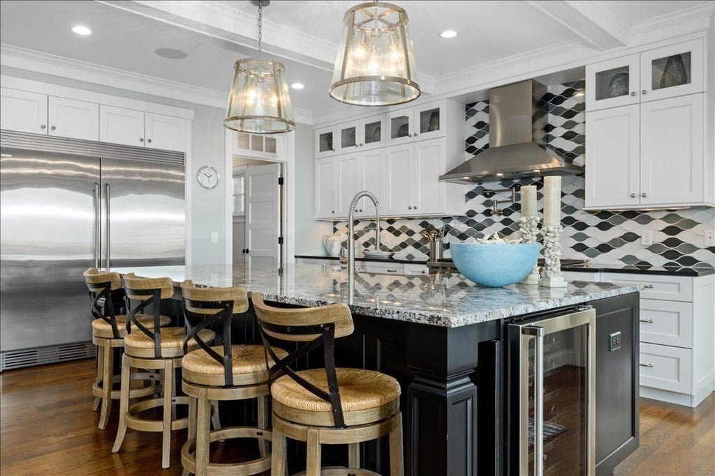 Cicular wood stools with backs are set up at an island with a black base. There is a wine refrigerator built into the side of the island and sink built into the counter. The stainless steel refrigerator is visible in the back left, and rows of white cabinets and the black-and-white tile backsplash are visible in the back left.