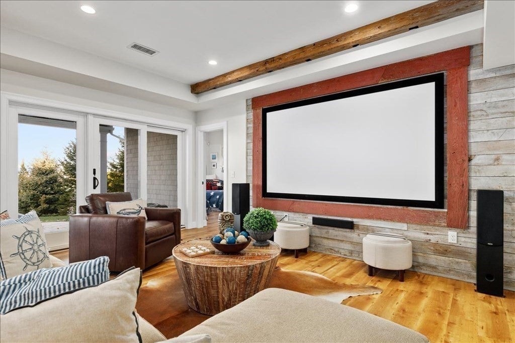 A large, white screen with a black border is attached to a wood plank wall. There a cushions of a couch visible in the foreground, and there sliding glass doors out to the back yard in the back left.