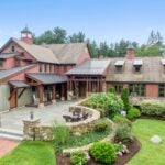 A view of the front of the home, which looks like a modern farmhouse that could be plopped right down in California wine country. The home has multiple roof lines. Rock walls and gardens circle away from the entrance. The home is a dark melon color with green-framed windows. a brick walkway leans up to two stone steps and to a stone walkway. The sun is shining with only a few clouds in the sky. The plantings are mature and the lawn is green.