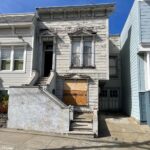 A dilapidated home has paint chipping all on the exterior. There are wood boards on the lower window.