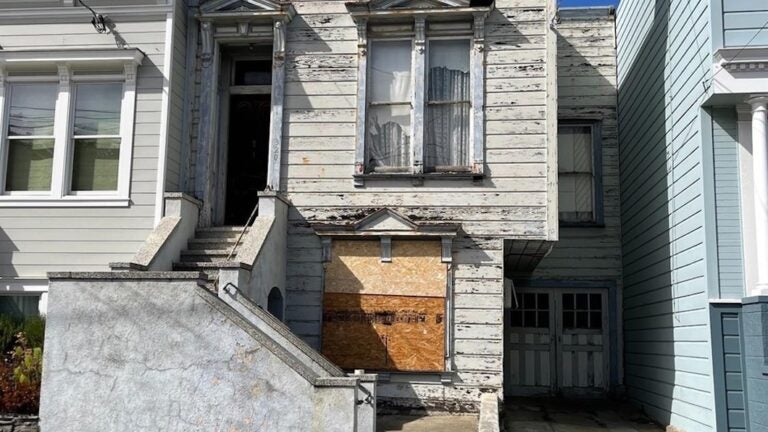 A dilapidated home has paint chipping all on the exterior. There are wood boards on the lower window.