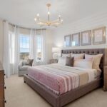 A bedroom with a king-size bed. The bed has an upholstered headboard that's brown. The walls are a soft white. An over-stuffed arm chair sits before a bay window with gauzy curtains. The floor is a cream-colored carpet. Four pictures in grayish-green frames hang over the bed in a row. A wide chandelier with candle-like lights hangs over the bed. A fraction of a wooden dresser is visible.