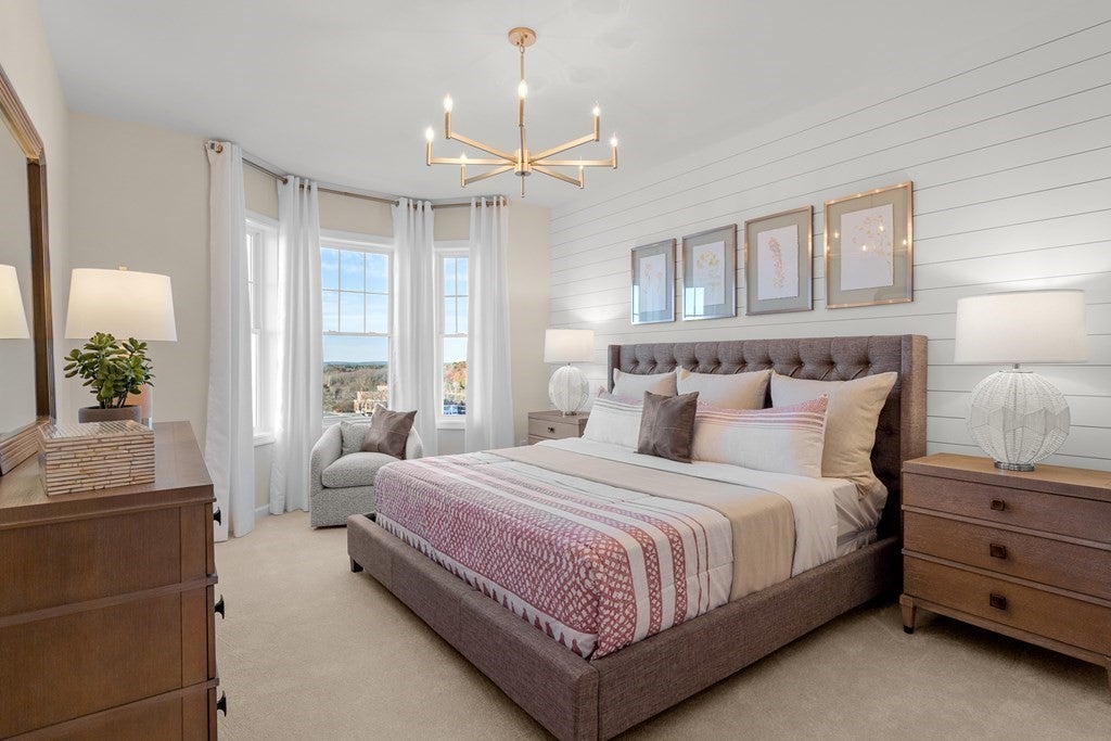 A bedroom with a king-size bed. The bed has an upholstered headboard that's brown. The walls are a soft white. An over-stuffed arm chair sits before a bay window with gauzy curtains. The floor is a cream-colored carpet. Four pictures in grayish-green frames hang over the bed in a row. A wide chandelier with candle-like lights hangs over the bed. A fraction of a wooden dresser is visible.