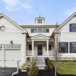 The exterior of the home has two pediments on each side, and a flat roof with a cupola sticking out in the middle. The front entrance is also in the middle, and that has a smaller pediment atop two columns. On the left of the entrance, there are two garage doors that look like barn or carriage doors. The home is white and has black shutters around the windows on the right side.