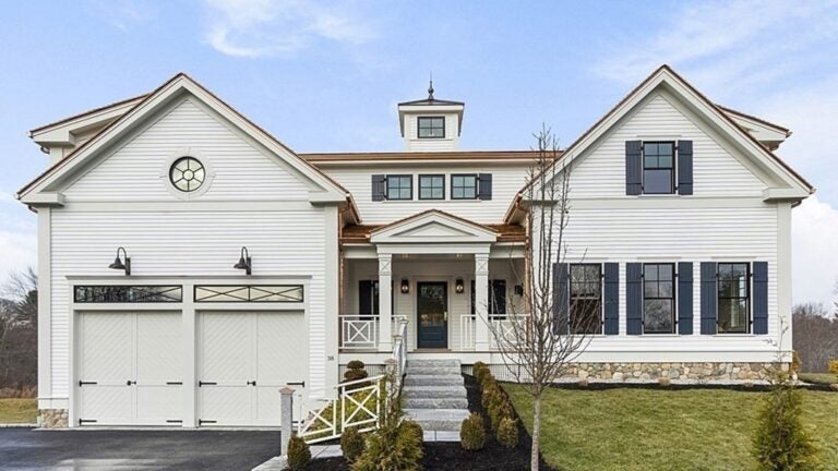 The exterior of the home has two pediments on each side, and a flat roof with a cupola sticking out in the middle. The front entrance is also in the middle, and that has a smaller pediment atop two columns. On the left of the entrance, there are two garage doors that look like barn or carriage doors. The home is white and has black shutters around the windows on the right side.