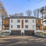 The exterior of this two unit townhouse is symmetrical, with brick walls leading up to the front doors of each unit on each of the far sides of the home. The exterior is mostly white, with wood panels on the sides where the house is set back slightly for the entrance. At the bottom of the home are two matching black garage doors with glass panels off to the right side of each.