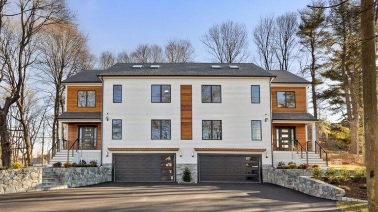 The exterior of this two unit townhouse is symmetrical, with brick walls leading up to the front doors of each unit on each of the far sides of the home. The exterior is mostly white, with wood panels on the sides where the house is set back slightly for the entrance. At the bottom of the home are two matching black garage doors with glass panels off to the right side of each.