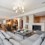 The living room features a gray sectional that defines the space and is bathed in natural light form a wall of black-framed windows. A candle-like chandelier hangs over the space. The rug is gray and white horizontal stripes. The space has a gas fireplace surrounded by board and batten wainscoting painted white.