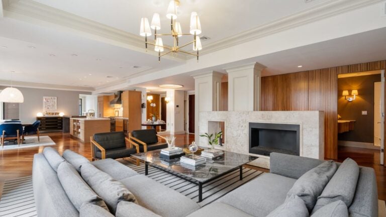 The living room features a gray sectional that defines the space and is bathed in natural light form a wall of black-framed windows. A candle-like chandelier hangs over the space. The rug is gray and white horizontal stripes. The space has a gas fireplace surrounded by board and batten wainscoting painted white.