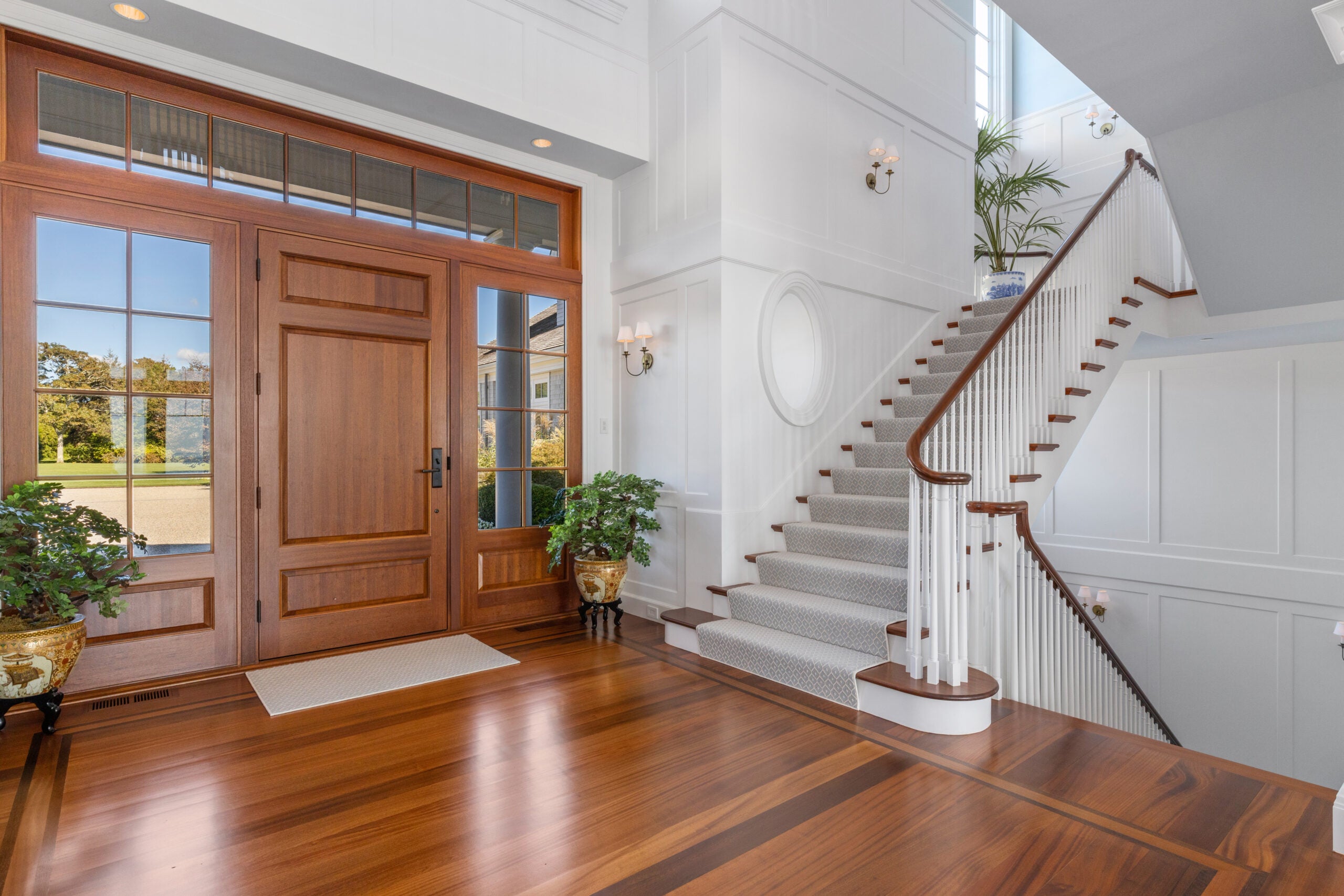 A view of a foyer with a wooden door with wide sidelights, board-and-batten wainscoting, wood stairs with a gray runner, sconces, and a hardwood floor with an inlay.