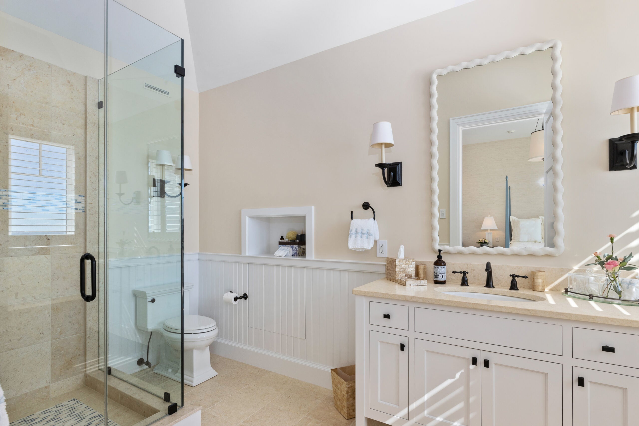 A bathroom with a single vanity, bead-board wainscoting, an inset shelf, a rectangular mirror with a white frame that mimics a frosted cake edge, white cabinetry, a single vanity with a sandy-colored top, and a frameless glass shower.
