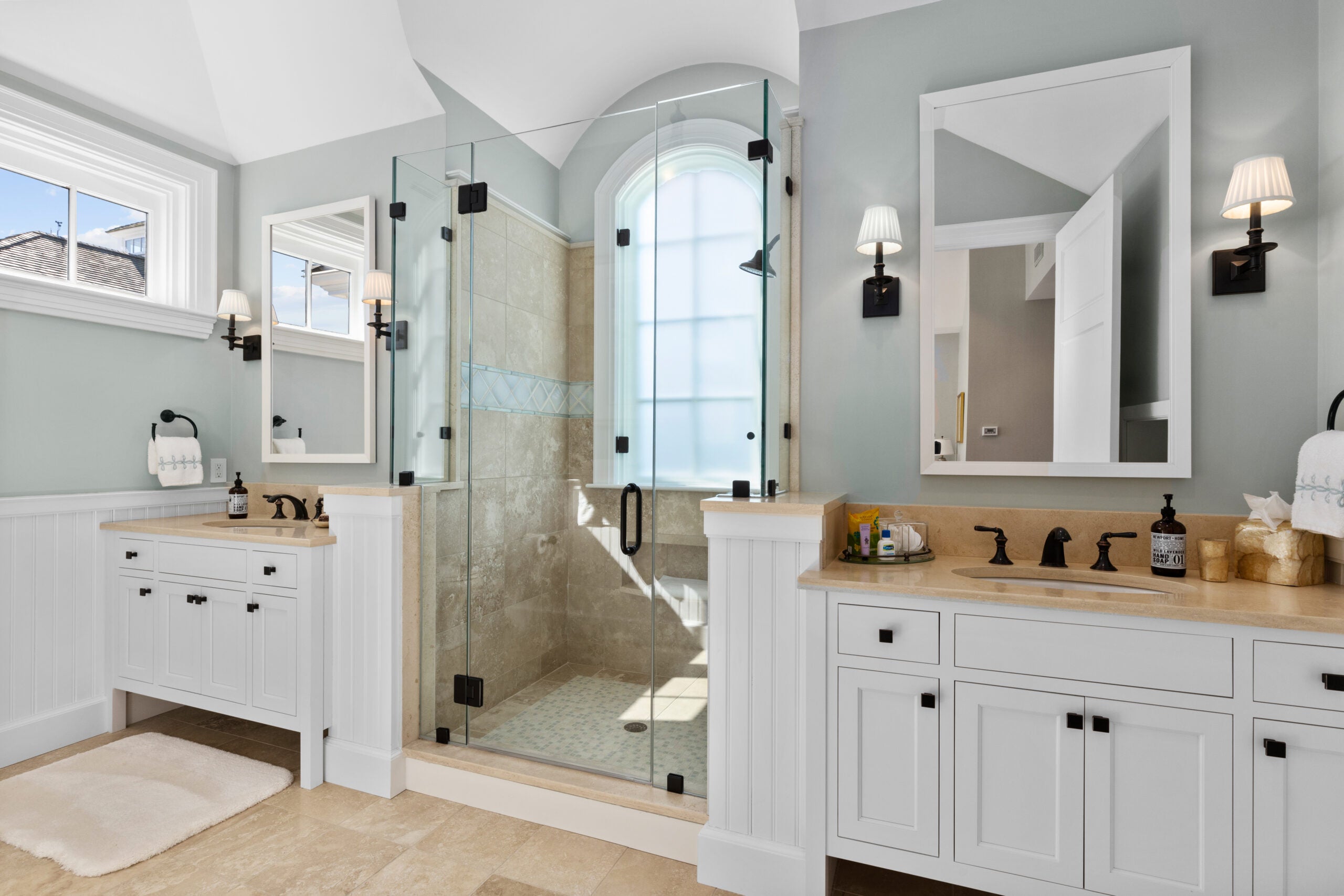 A shower with an arched window, black fixtures, and a ceramic surround with an inlay is flanked by two vanities with black fixtures, tall and rectangular white-framed mirrors, shaded sconces, white cabinetry with black handles, and sandy-colored counters. The flooring is a sand-colored tile, and the walls are gray.