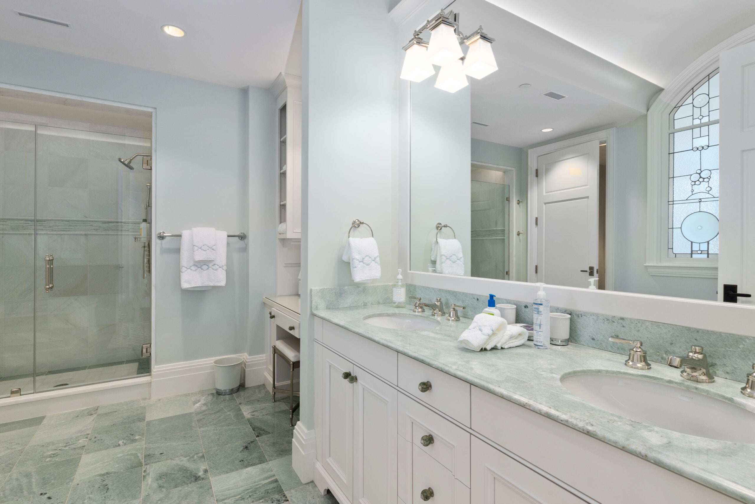 A bathroom with a curbed shower with glass doors, recessed lighting dual sinks, a greenish-gray counter, white cabinetry, a dressing table, and greenish-gray tile. a stained-glass window depicting bubbles is partially visible.