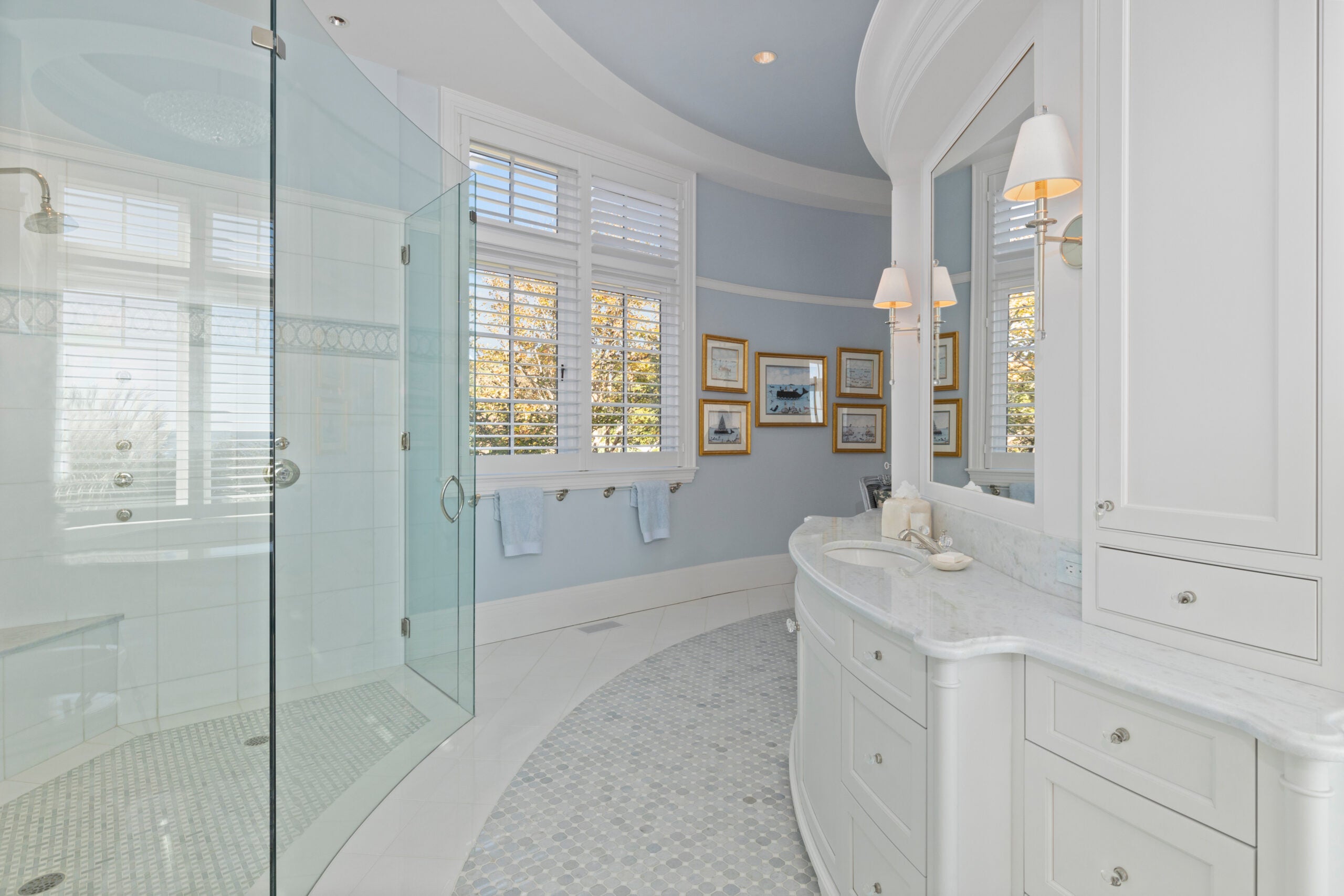 A curvy bathroom with white cabinetry, a frameless glass shower, and blue walls. A pair of windows floods the space with light. A small overall sink in a marble-veined vanity is visible. The walls are blue and lined with gold-framed pictures. The flooring is a mix of marble tile and small gray circular tile.