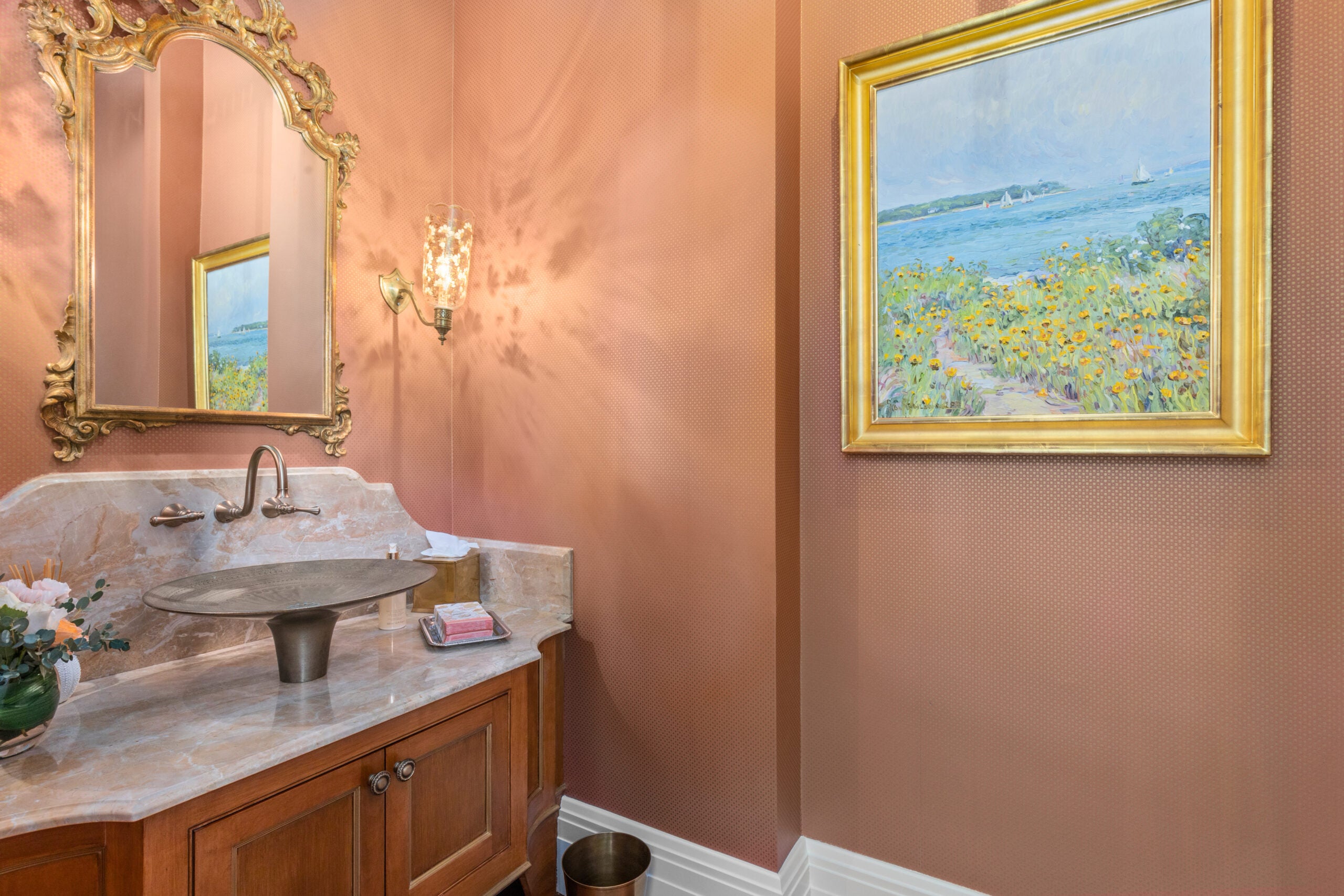 A powder room with a small-pattered wallpaper the color of terra cotta, a gold-framed mirror, a gold-framed picture of a seascape, and a vanity with wood cabinetry, a wall-mounted faucet, and a vessel sink.