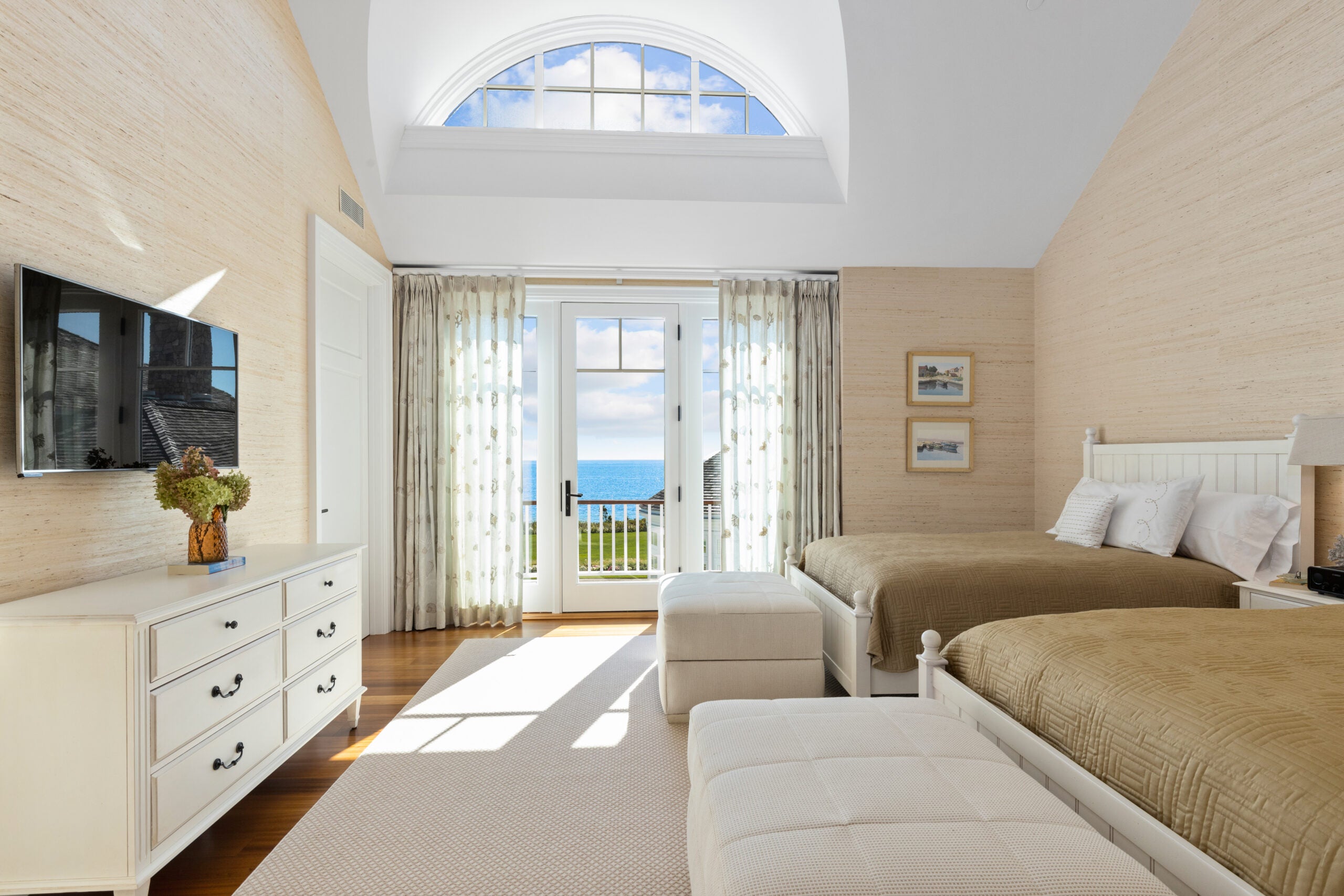 A bedroom with two matching wooden king-size beds. An overstuffed ottoman sits at the end of each bed. The bed linens are a light gold, which matches the grasscloth wallpaper. A slider with gauzy floral drapes partially covers a glass door to a deck overlooking the ocean and a section of the house. An muntined oval window is inset into the ceiling. A TV hangs over a white dresser, and the hardwood floor is covered in a white rug.