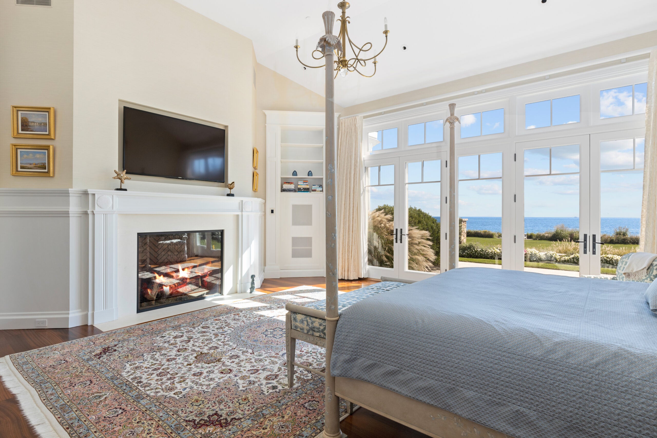 A four-poster bed with blue linens sits before three glass doors to a patio, as well as a gas fireplace framed in white. An Oriental rugs sits between the fireplace and the bed.