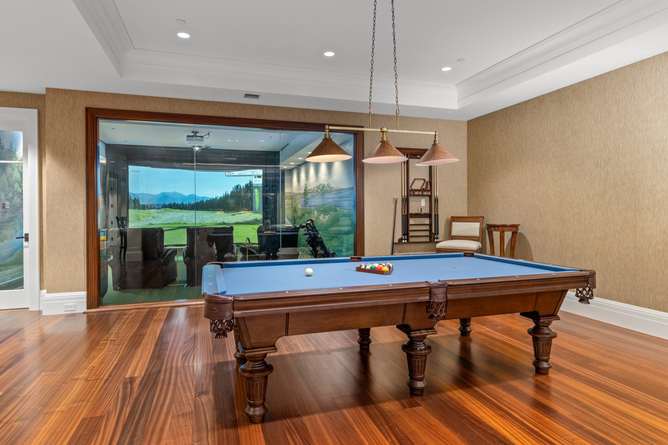 A blue billiards table sits under a bank of three iron-hooded lights. The golf simulation room sits behind glass in the background. The pool table is under a tray ceiling with recessed lighting, and the walls are papered in a sandy-brown glasscloth. The floor is hardwood.