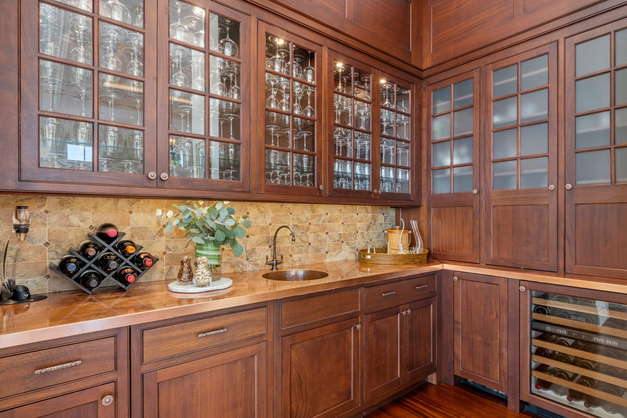 The butler's pantry has a mix of dark wood cabinetry with wood and glass (smokey or clear) doors. The countertop is glossy and bronze-colored. There is a small oval sink, myriad wine glasses behind the clear-glass doors, a sandy-colored tile backsplash with inlays, and a wine chiller.