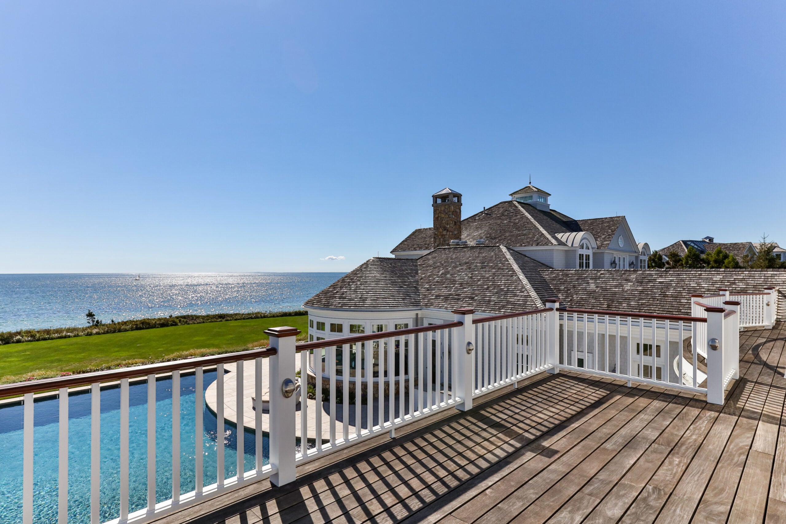 A wood deck with white balusters and dark wood railings looks out over the pool, the lawn, and to the sea. The sky is nearly cloudless. Off to the right, there is one of two homes on the property.