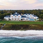 An aerial view of the home from the ocean. Waves lap at a sandy beach. Off to the left there are stairs to a grassy lawn. A wooded expanses behind the estate shows hints of other homes.