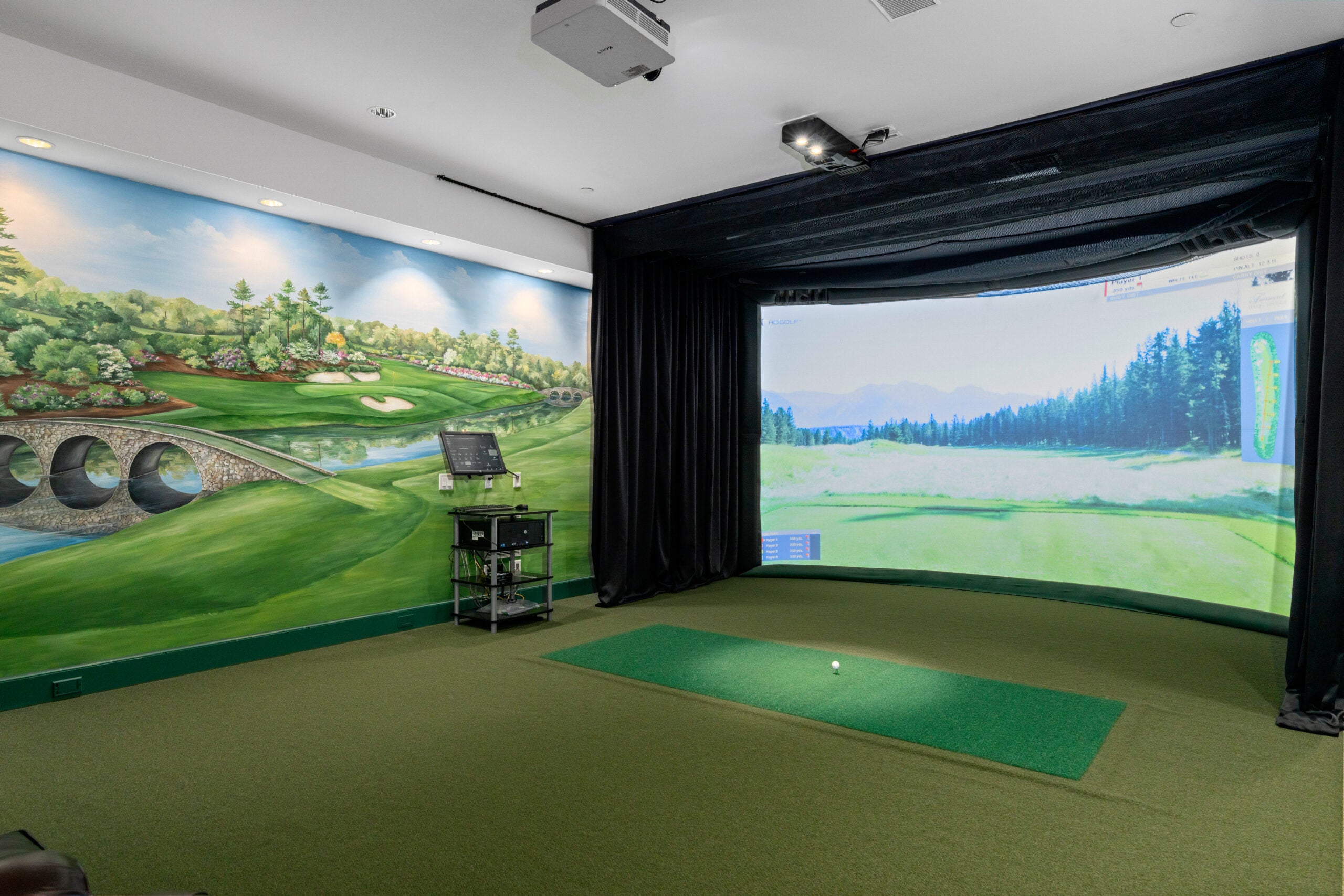 A golf simulator room with the projection of a fairway on the screen. The flooring is a grass-colored carpet. There is painted mural of a golf hole off the left.