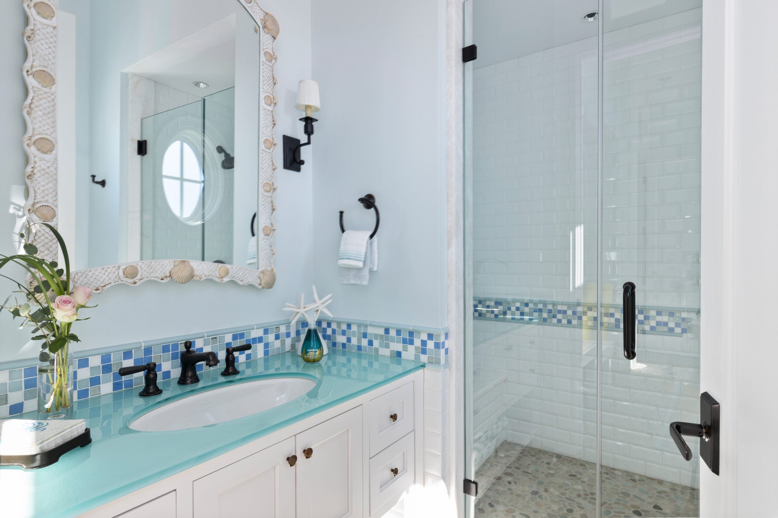 A bathroom with a green, white, and blue glass tile inlay that is mimicked in the shower. A seashell mirror hangs over a sink with an aqua glass top. The cabinetry is white. The shower has a pebblestone floor.