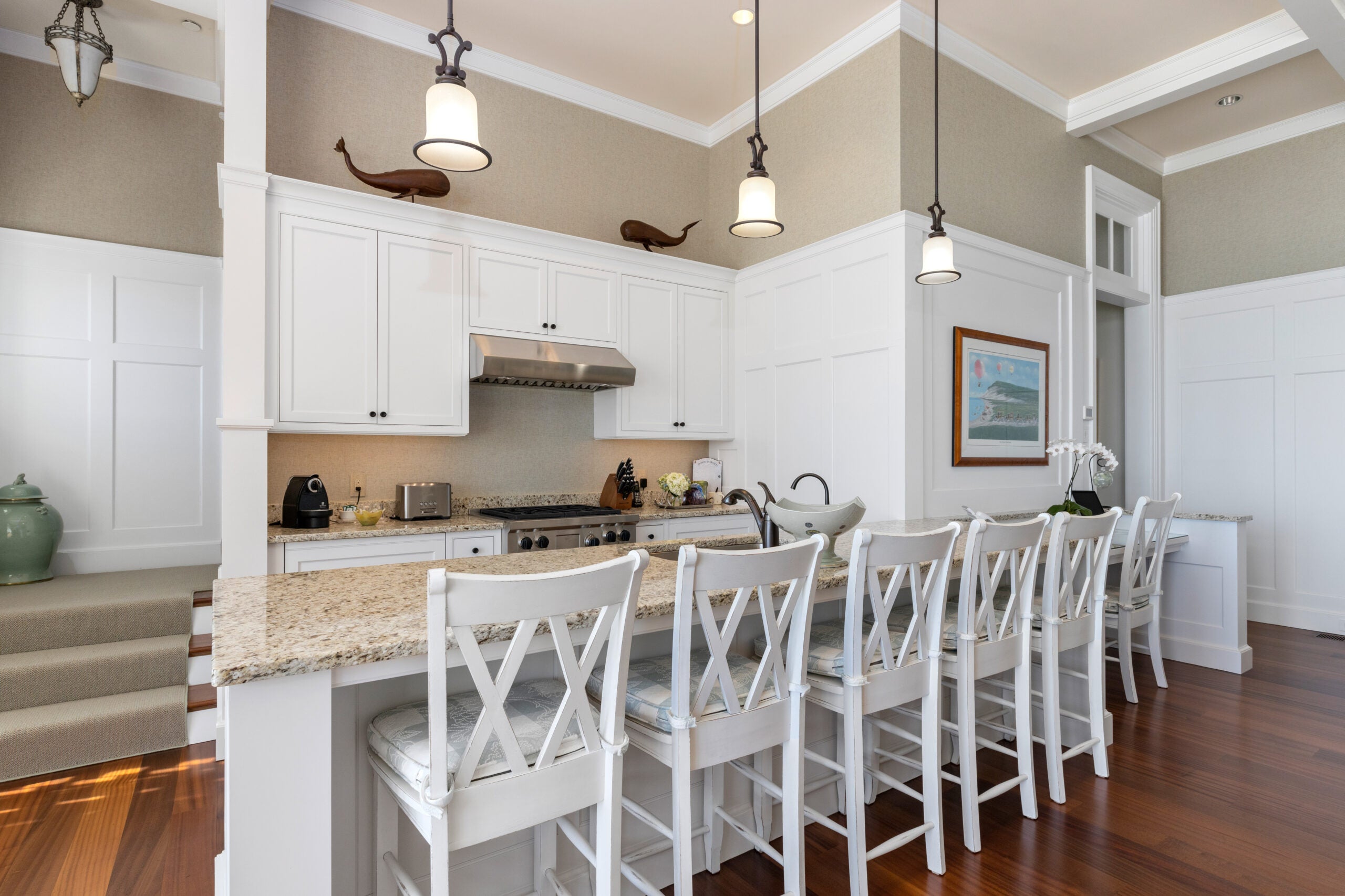 The guest house kitchen, which has a long granite-topped counter with seating for six, white raised-panel cabinetry, crown molding, three pendant lights, recessed lighting, beige walls, hardwood flooring, and board-and-batten wainscoting.