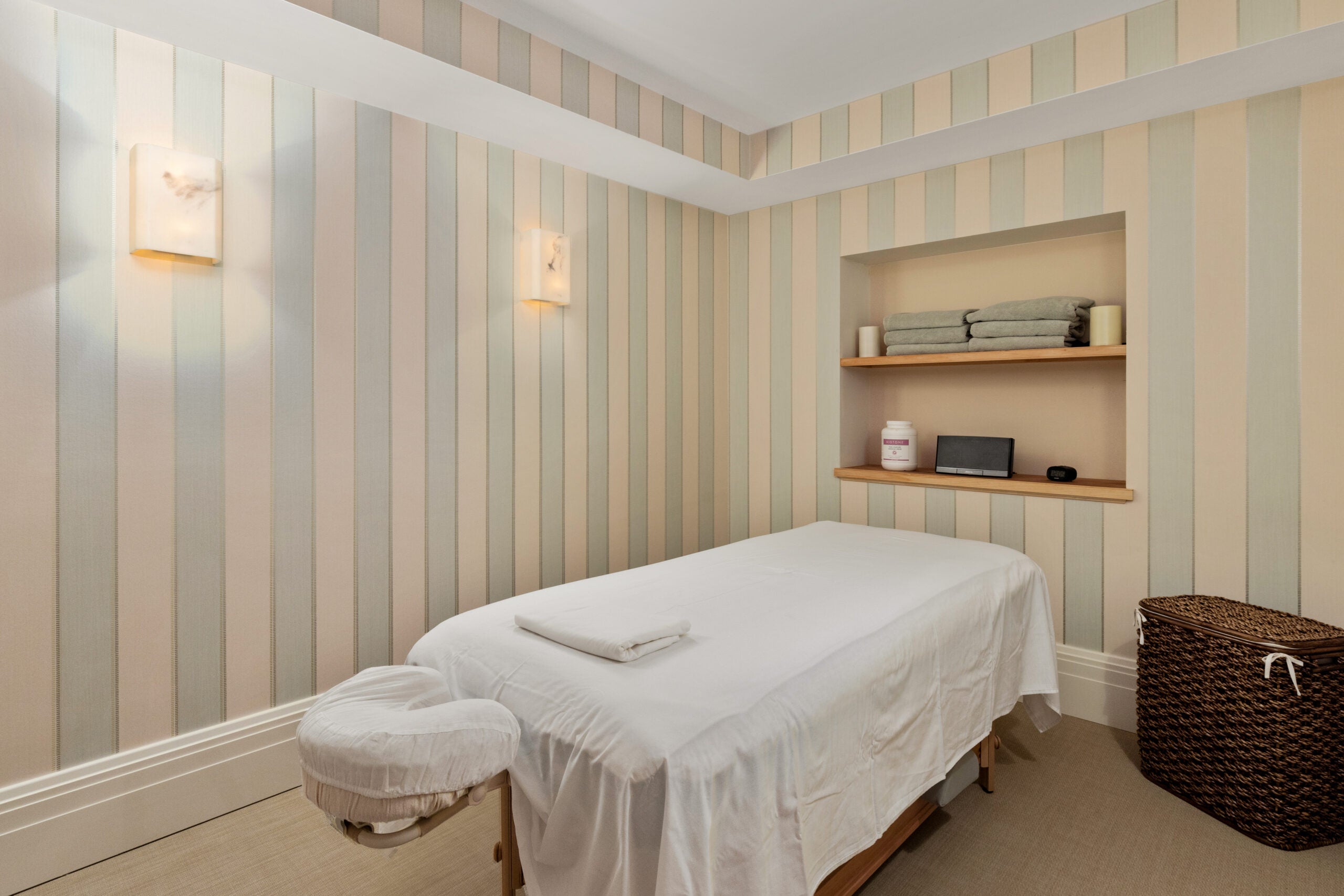 The massage room, which has box sconces, a massage table with white linen, a whicker basket, and shelves holding white candles and green towels. The wallpaper in this space is green and cream stripes. The floor is carpeted.