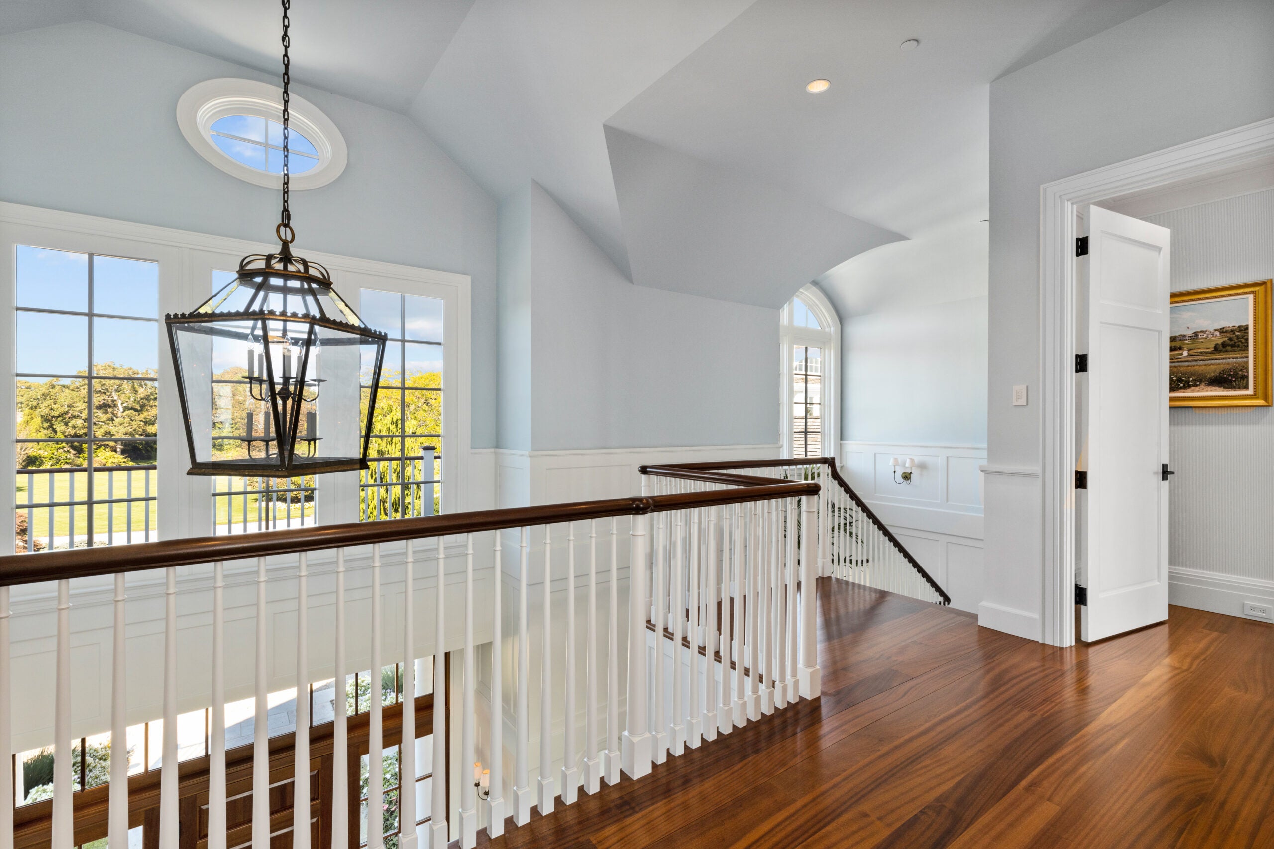 The landing off the top of the stairwell shows a hardwood floor with lots of swirls and a high gloss. A big lantern hangs over the space. The balusters are myriad and white, but the railings are a dark wood. The railing looks over to an oval window and an expansive three-unit window overlooking a deck and a yard with numerous trees. The walls of this space are gray and there is board-and-batten wainscoting lining the stairway.