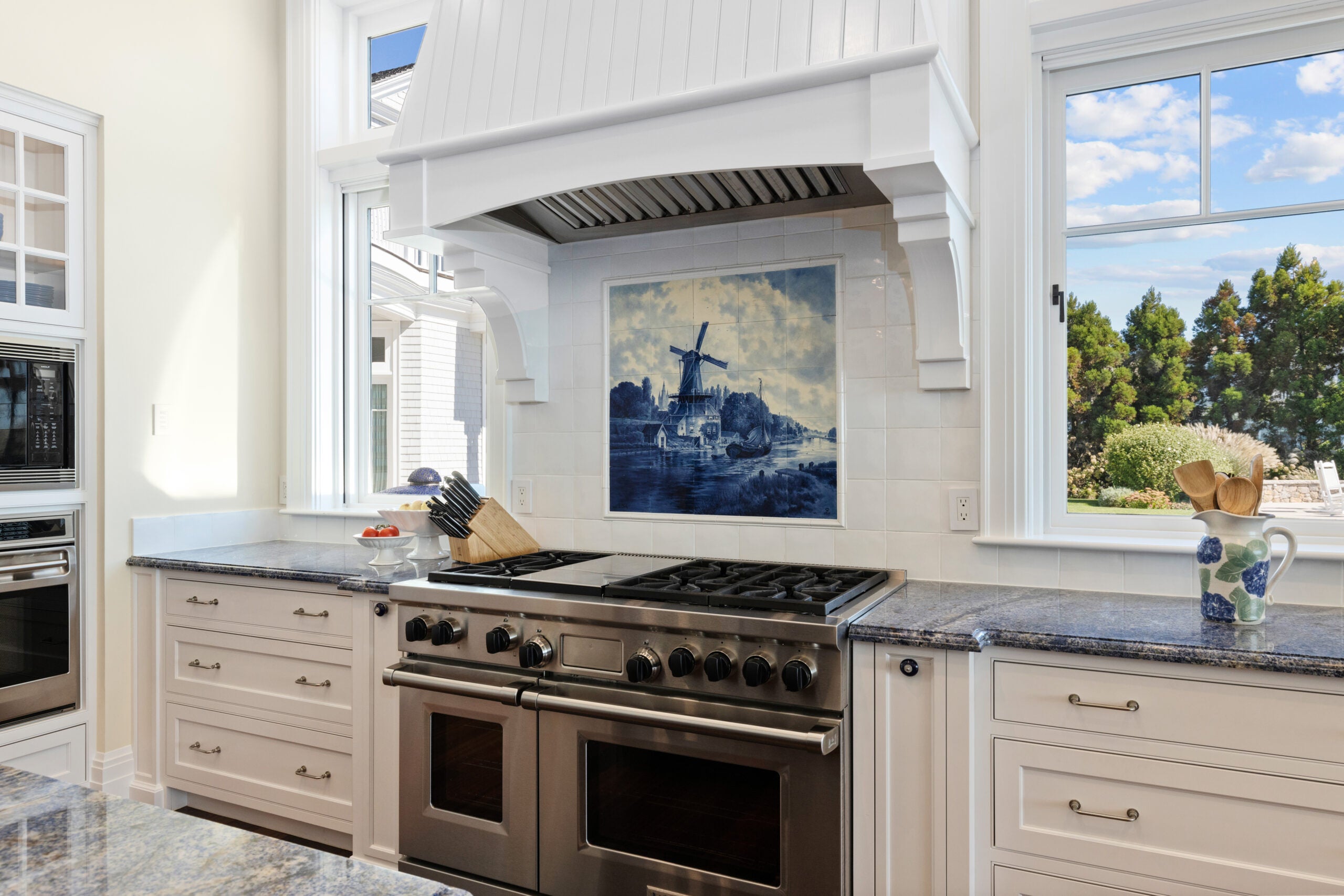 A detail shot of the kithen counter, which is blue and a double oven gas stove that is flanked by two 2-over-1 windows offering a view of the backyard. It is a sunny day with a few clouds, and the yard has mature trees whose low height is common along the coastline. The bushes are a mixture of grasses. The backsplash behind the stove is a white tile with a blue inlay of a windmill scene. There is at least one other oven off to the left, and the cabinetry that flanks the stove is composed of drawers. The stove hood is wood painted white.